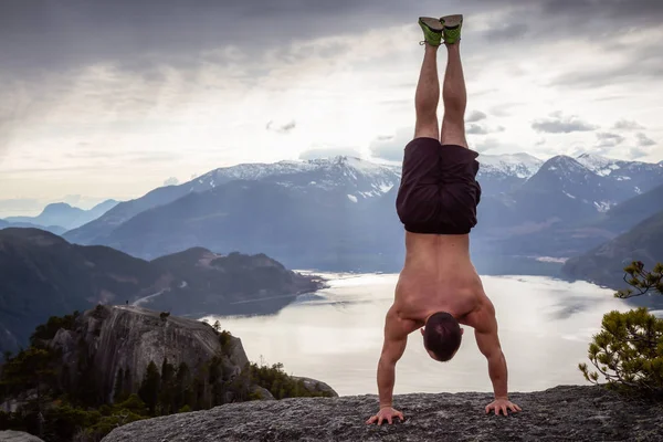 Fit and Muscular Young Man is doing a handstand on top of the Mountain during a cloudy day. Taken on Chief Mountain in Squamish, North of Vancouver, BC, Canada.