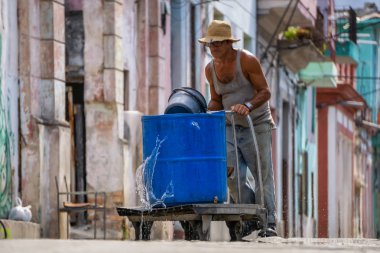 Havana, Küba - 17 Mayıs 2019: Yaşlı adam sıcak güneşli bir günde Eski Havana Şehrinin sokaklarında su dolu bir konteyner itiyor.