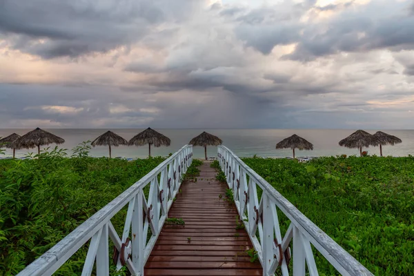 Beautiful view of a wooden path leading to the sandy beach on the Caribbean Sea in Cuba during a cloudy and rainy morning sunrise.