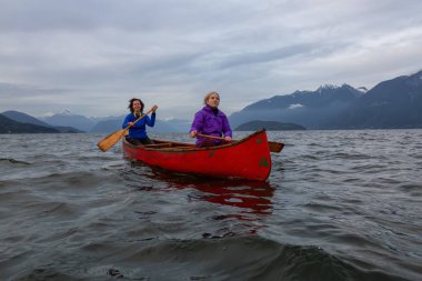 Kırmızı kano üzerinde çift maceracı kadın arkadaşlar bulutlu bir gün batımı sırasında Howe Sound kürek vardır. Horseshoe Bay, Batı Vancouver, Bc, Kanada.