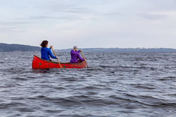 在多云的日落期间 一对冒险的女性朋友在一条红色的独木舟上在豪湾划船 位于加拿大不列颠哥伦比亚省温哥华以西的博文岛附近 — 图库照片