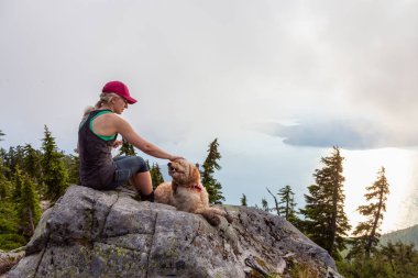 Adventurous Girl is hiking with a dog on top of Unnecessary Mountain during a sunny and cloudy summer day. Located in West Vancouver, British Columbia, Canada. clipart