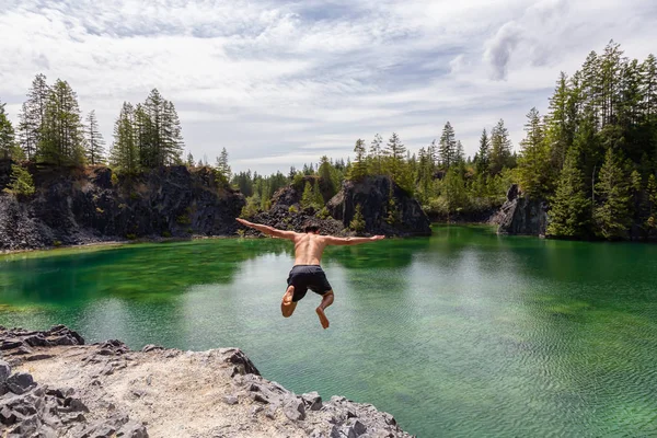 Athletic and Adventurous Man is Cliff Jumping into a Green Colored Glacier Lake during a hot and sunny summer day. Taken in British Columbia, Canada.