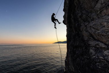 Güneşli bir yaz gün batımı sırasında rocly okyanus kıyısında dik bir uçurumdan aşağı rappelling tanınmayan bir adam silueti. Lighthouse Park, Batı Vancouver, British Columbia, Kanada'da çekildi.