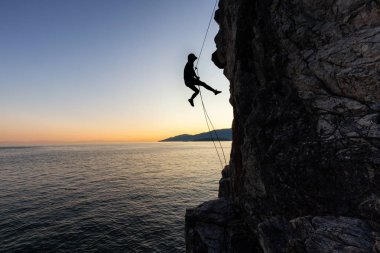 Güneşli bir yaz gün batımı sırasında rocly okyanus kıyısında dik bir uçurumdan aşağı rappelling tanınmayan bir adam silueti. Lighthouse Park, Batı Vancouver, British Columbia, Kanada'da çekildi.
