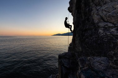 Güneşli bir yaz gün batımı sırasında rocly okyanus kıyısında dik bir uçurumdan aşağı rappelling tanınmayan bir adam silueti. Lighthouse Park, Batı Vancouver, British Columbia, Kanada'da çekildi.