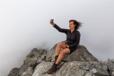 Genç Maceracı Kız bulutlu bir yaz sabahı bir selfie alarak engebeli kayalık dağın tepesinde. Crown Mountain'da çekilmiş, Kuzey Vancouver, Bc, Kanada.