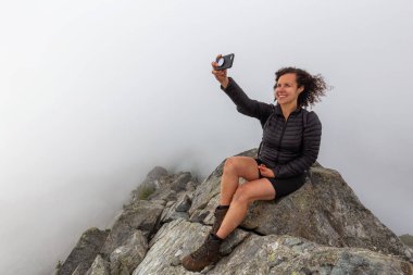 Genç Maceracı Kız bulutlu bir yaz sabahı bir selfie alarak engebeli kayalık dağın tepesinde. Crown Mountain'da çekilmiş, Kuzey Vancouver, Bc, Kanada.