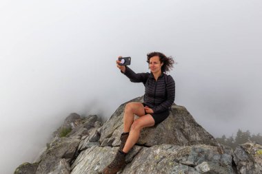 Genç Maceracı Kız bulutlu bir yaz sabahı bir selfie alarak engebeli kayalık dağın tepesinde. Crown Mountain'da çekilmiş, Kuzey Vancouver, Bc, Kanada.