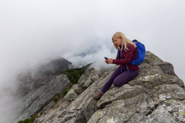 Genç Maceracı Kız bulutlu bir yaz sabahı bir selfie alarak engebeli kayalık dağın tepesinde. Crown Mountain'da çekilmiş, Kuzey Vancouver, Bc, Kanada.