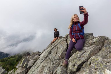 Genç Maceracı Kız bulutlu bir yaz sabahı bir selfie alarak engebeli kayalık dağın tepesinde. Crown Mountain'da çekilmiş, Kuzey Vancouver, Bc, Kanada.