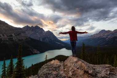 Adventurous girl with open arms standing on the edge of a cliff overlooking the beautiful Canadian Rockies and Peyto Lake during a vibrant summer sunset. Taken in Banff National Park, Alberta, Canada.