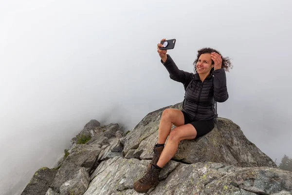 Genç Maceracı Kız bulutlu bir yaz sabahı bir selfie alarak engebeli kayalık dağın tepesinde. Crown Mountain'da çekilmiş, Kuzey Vancouver, Bc, Kanada.