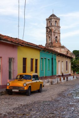 Trinidad, Küba - 12 Haziran 2019: Canlı güneşli bir gün batımında arka planda Kilise ile küçük bir Küba Kasabası sokaklarında eski bir Klasik Taksi Arabası Görünümü.