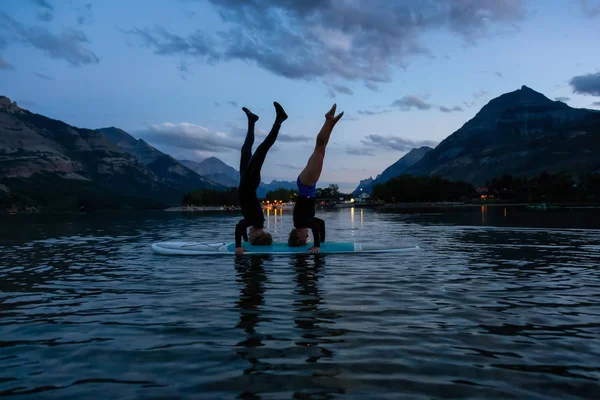 Waterton Lakes Ulusal Parkı, Alberta, Kanada - 31 Ağustos 2019: Maceracı Kız canlı bir yaz gün batımı nda bir kürek tahtası üzerinde bir baş standı yapıyor.