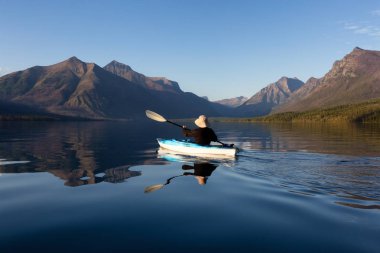 Adventurous Man Kayaking in Lake McDonald during a sunny summer evening with American Rocky Mountains in the background. Taken in Glacier National Park, Montana, USA. clipart