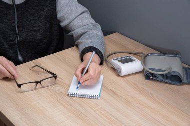 Kadın evde kan basıncını bağımsız olarak ölçer ve ölçümleri bir deftere kaydeder.