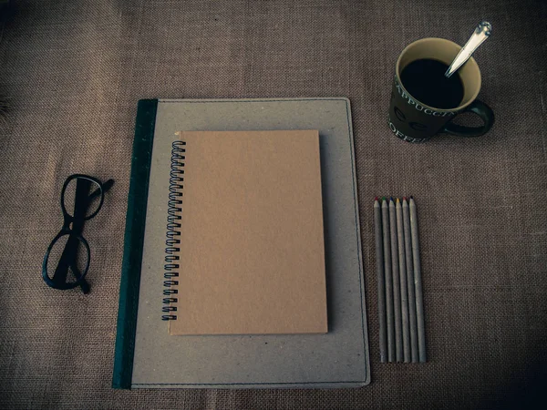 Vintage style. Organized desk with binder, closed notebook, eye glasses, one cup of coffee, pencils and burlap background