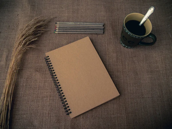 Vintage style. Organized desk with closed notebook, dry grass, one cup of coffee, pencils and burlap background