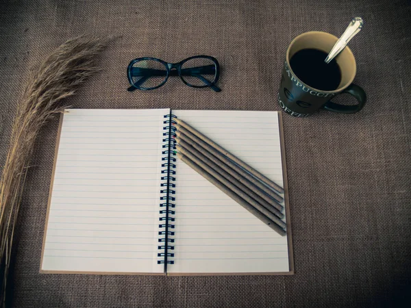 Vintage style. Organized desk with open notebook, dry grass, one cup of coffee, eye glasses, pencils and burlap background