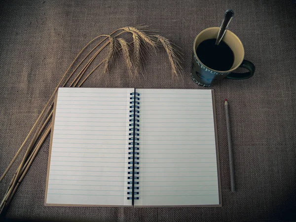 Vintage style. Organized desk with open notebook, a cup of coffee, dry grass, pencils and burlap background