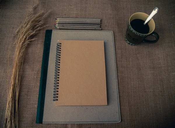 Vintage style. Organized desk with binder, closed notebook, dry grass, one cup of coffee, pencils and burlap background