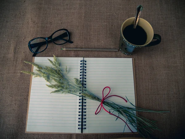 Vintage style. Organized desk with open notebook, a cup of coffee, dry grass, eye glasses, pencils and burlap background
