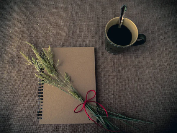 Vintage style. Organized desk with closed notebook, a cup of coffee, dry grass, pencils and burlap background