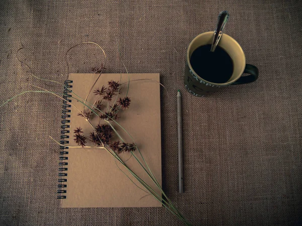 Vintage style. Organized desk with closed notebook, dry grass, one cup of coffee, pencils and burlap background