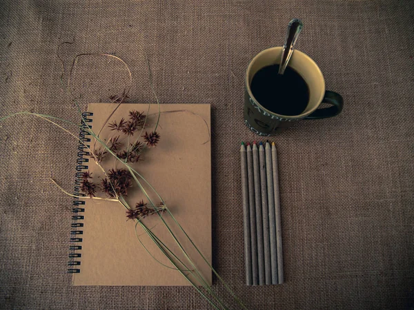 Vintage style. Organized desk with closed notebook, dry grass, one cup of coffee, pencils and burlap background