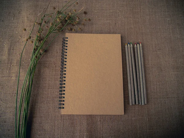 Vintage style. Organized desk with closed notebook, dry grass, pencils and burlap background