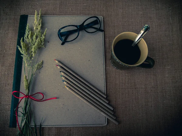 Vintage style. Organized desk with binder, eye glasses, a cup of coffee, dry grass, pencils and burlap background