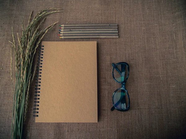 Vintage style. Organized desk with closed notebook, eye glasses, dry grass, pencils and burlap background