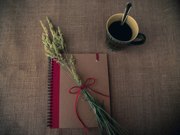 Vintage style. Organized desk with closed notebook, dry grass, one cup of coffee and burlap background