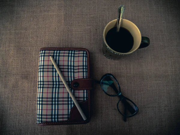 Vintage style. Organized desk with closed diary, eye glasses, a cup of coffee, pencil and burlap background