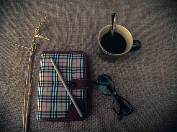 Vintage style. Organized desk with closed diary, eye glasses, a cup of coffee, dry grass, pencil and burlap background