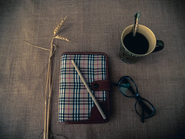 Vintage style. Organized desk with closed diary, eye glasses, a cup of coffee, dry grass, pencil and burlap background