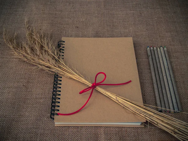 Vintage style. Organized desk with closed notebook, dry grass, pencils and burlap background