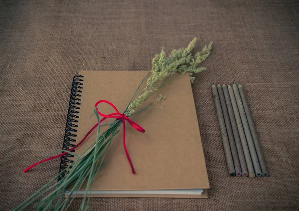 Vintage style. Organized desk with closed notebook, dry grass, pencils and burlap background