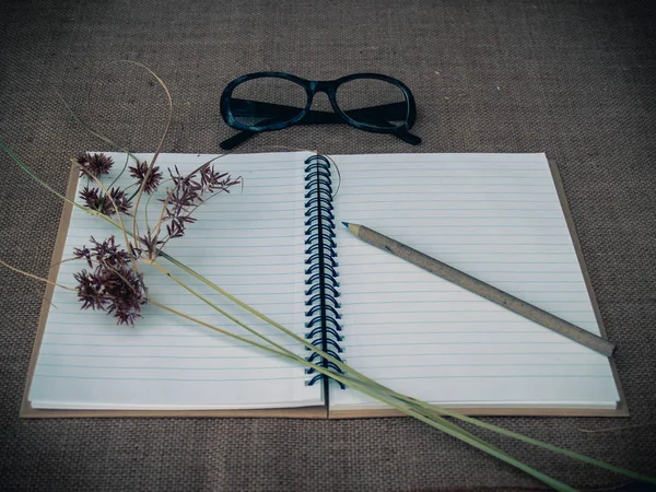 Vintage style. Organized desk with open notebook, eye glasses, dry grass, pencils and burlap background