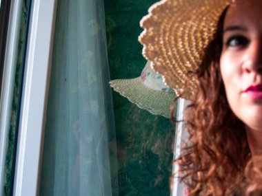 Kırsal bir evde bir pencereden gelen doğal ışık ile hasır şapka kıvırcık saçlı Genç bir kadın, bir pencerede yansıması