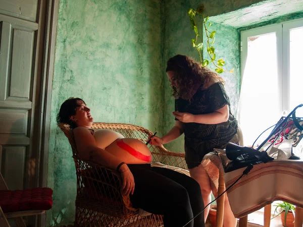 Female artist painting the belly of a pregnant young woman with body paints and brushes