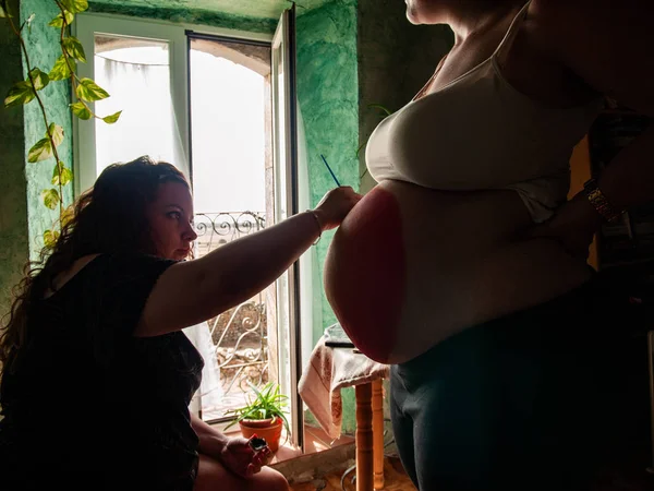 Female artist painting the belly of a pregnant young woman with body paints and brushes