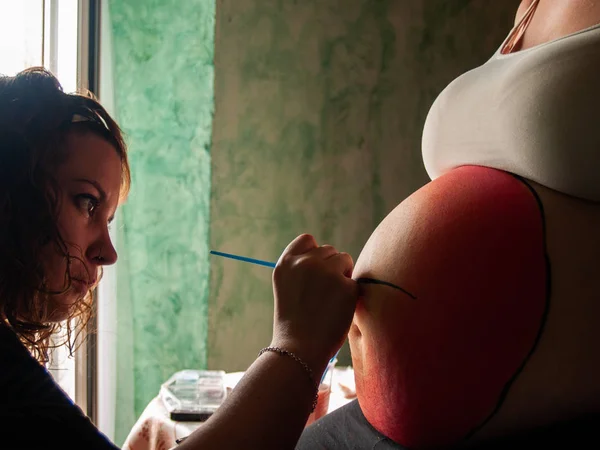 Female artist painting the belly of a pregnant young woman with body paints and brushes