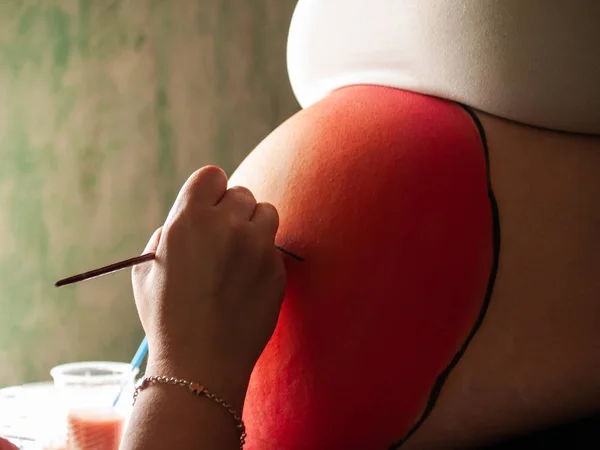 Female artist painting the belly of a pregnant young woman with body paints and brushes