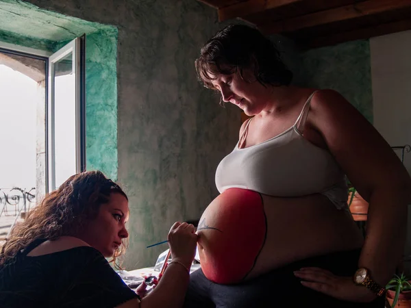 Female artist painting the belly of a pregnant young woman with body paints and brushes