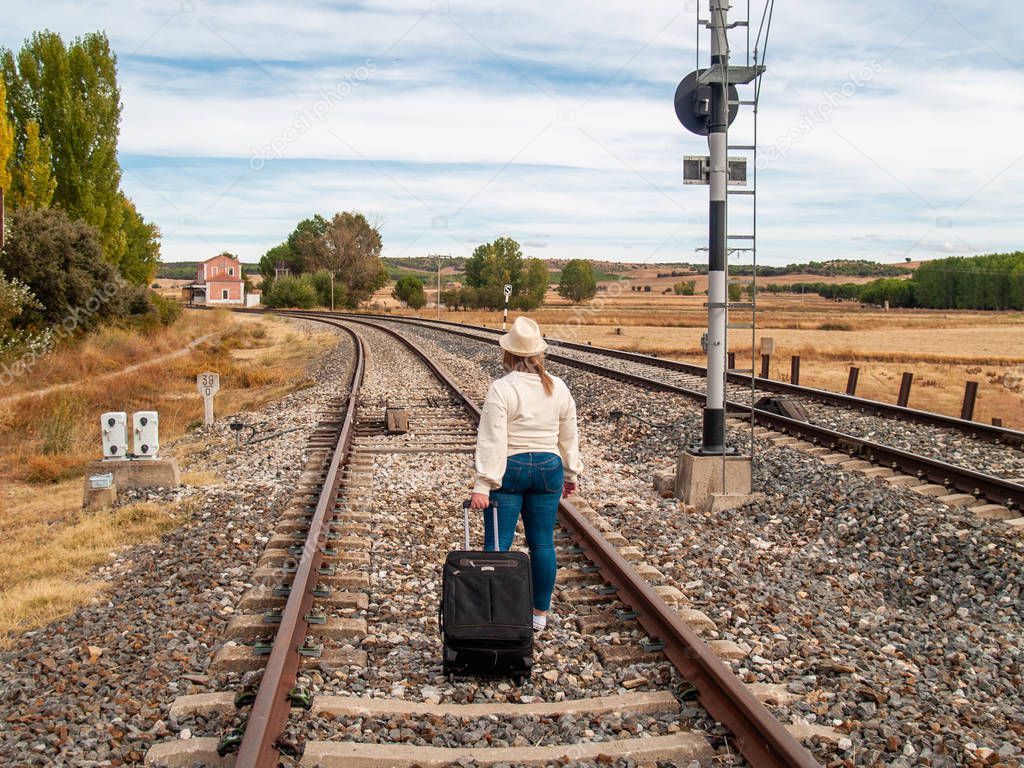A sad blonde woman with a straw hat walking on the train tracks carrying a suitcase in autumn