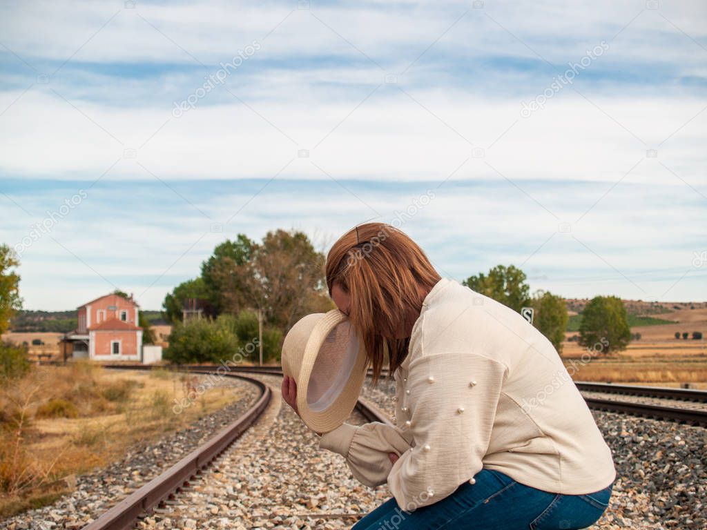 A sad blonde woman with a straw hat on the train tracks sitting on a suitcase in autumn