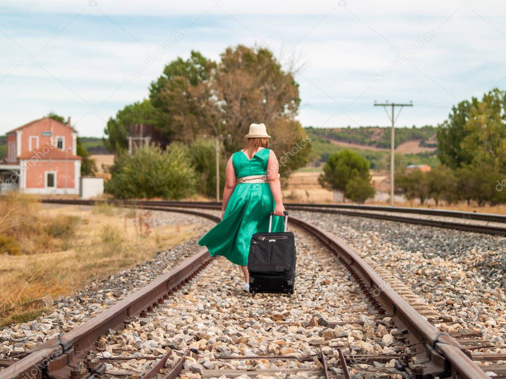 A blonde woman with a green dress and a straw hat walking on the train tracks carrying a suitcase in autumn