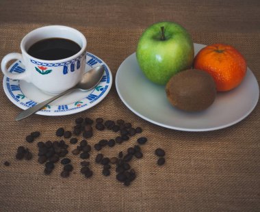 Porselen kahve fincan, kahve çekirdekleri, eski bir kaşık ve bir tabak bir yeşil elma, bir mandalina ve vintage çuval bezi arka plan üzerinde bir kivi ile kahvaltı